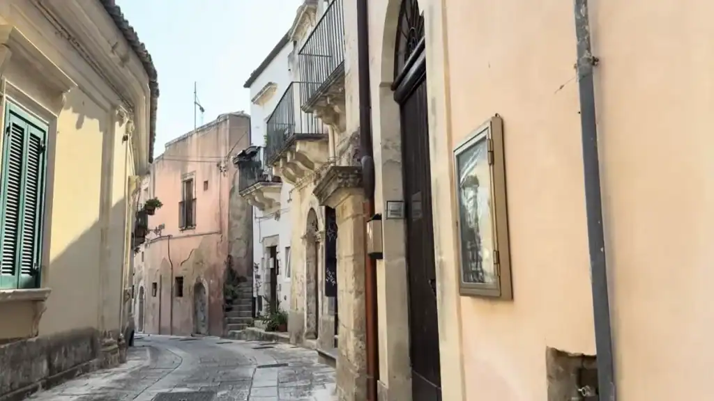 Stone steps climb between worn pastel houses along one of the quieter Ragusa Ibla streets, carved baroque doorframes interrupting the otherwise plain residential walls