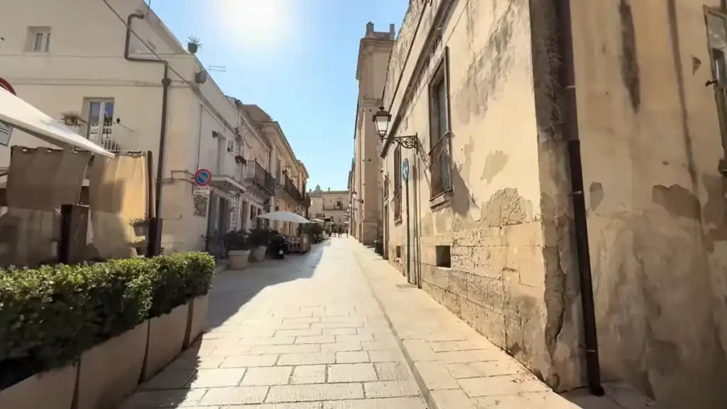 Narrow Ragusa Ibla streets lined with weathered limestone buildings, a café terrace on the left and a church tower visible at the far end