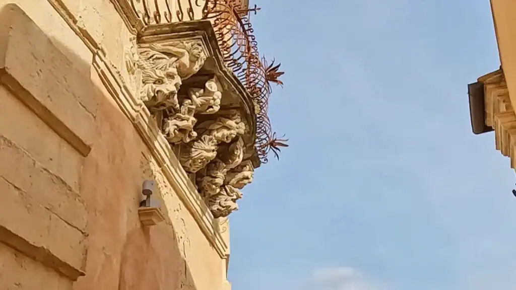 Grotesque carved figures supporting a wrought-iron balcony bracket on a baroque palazzo facade in Ragusa Ibla, mouths open, stacked in dense relief