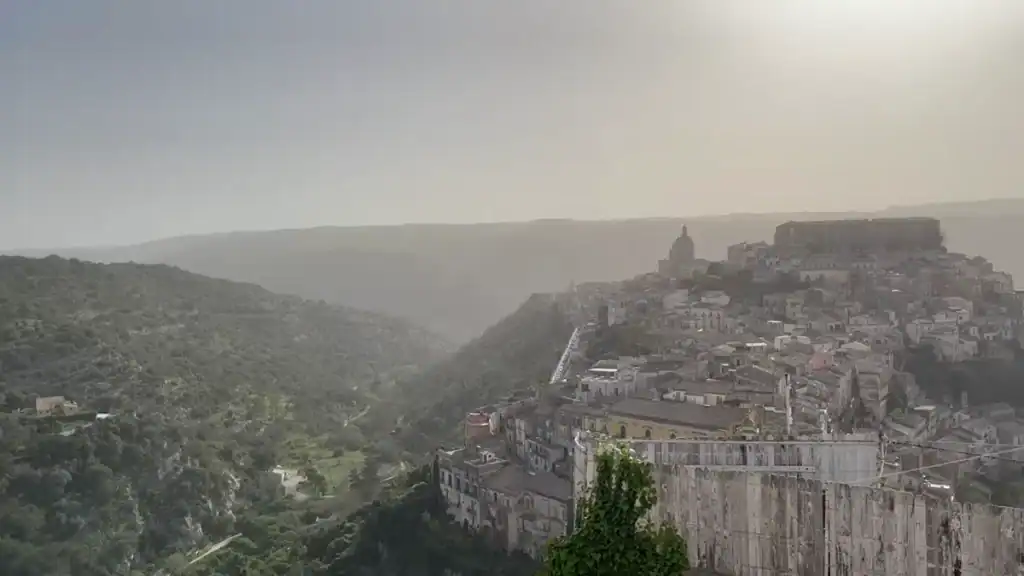 Ragusa Ibla occupying a ridge on the right, its dome and rooftops fading into hazy air above a deep wooded gorge.