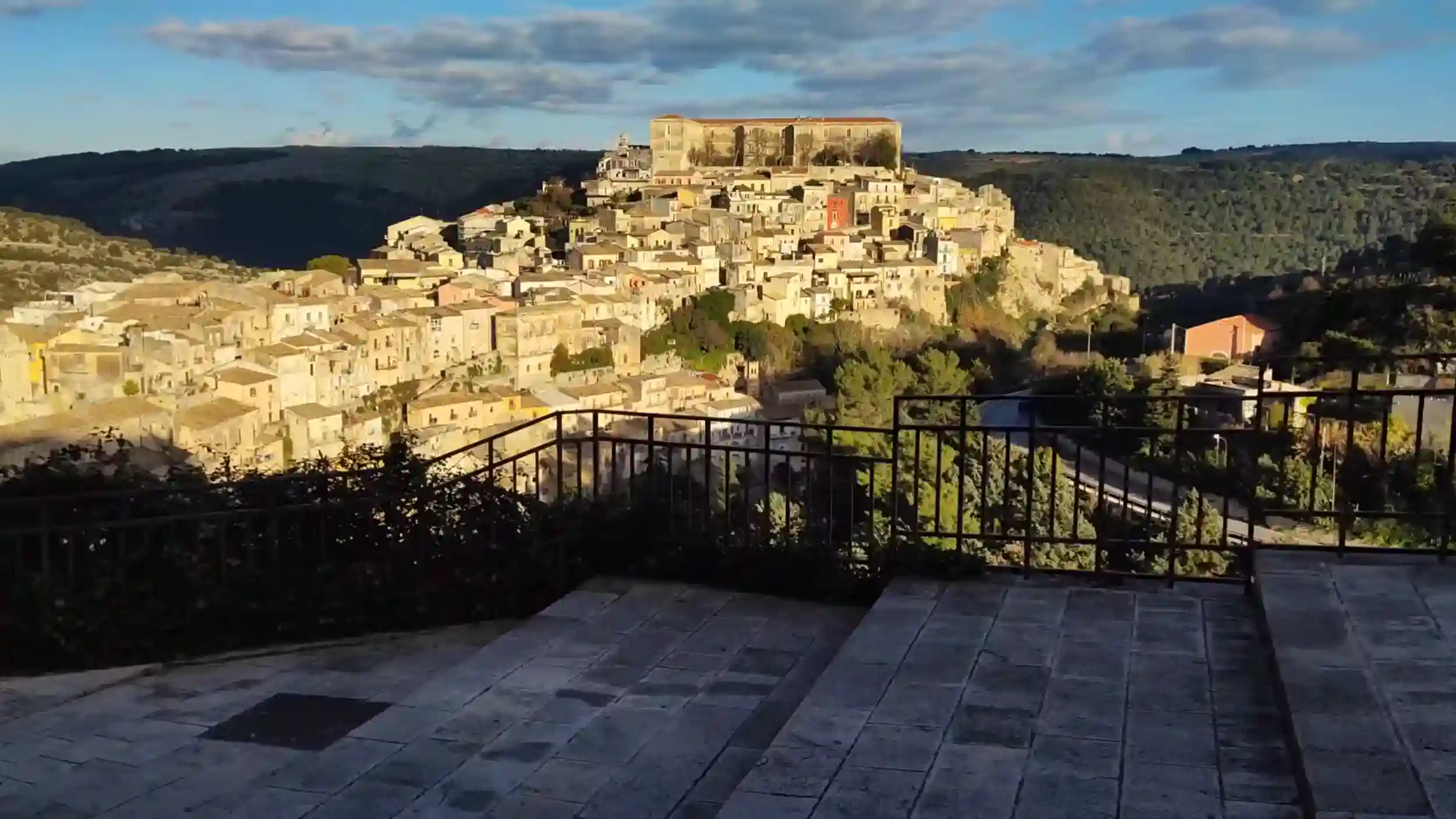 Ragusa Ibla stacked across a hilltop, its honey-coloured buildings rising to a large palazzo at the summit, seen from a paved terrace with iron railings - walking in Ragusa reveals approaches like this one across the valley