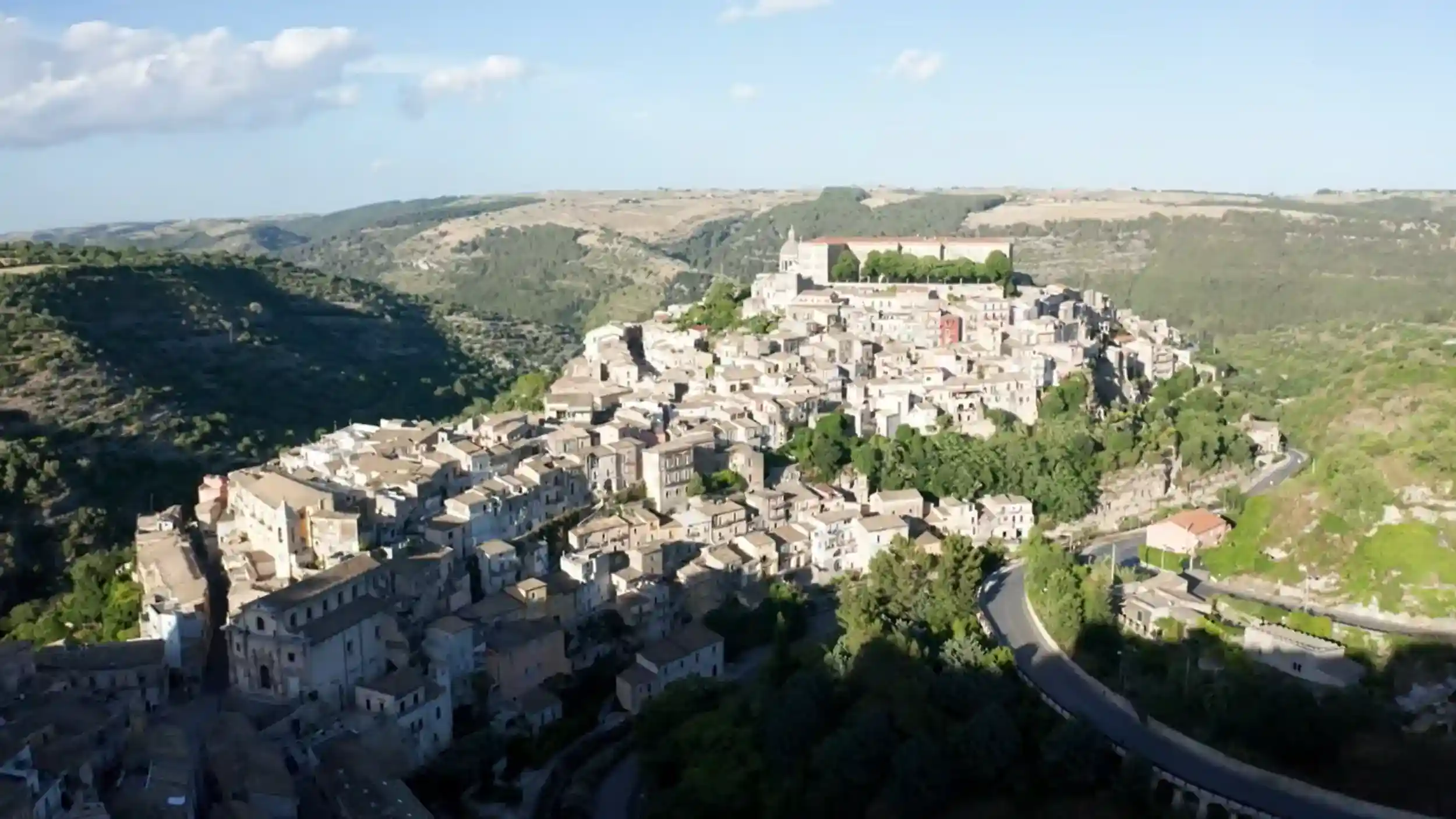 Ragusa Ibla spread across a rocky ridge, its stone buildings stacked densely downhill with a road curving through the valley below - seen on a drive to Ragusa through the surrounding green hills.