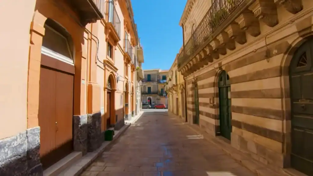 Narrow street in Ragusa Superiore with ochre-painted facades on the left and a striped stone building with arched doorways on the right, opening onto a small piazza ahead