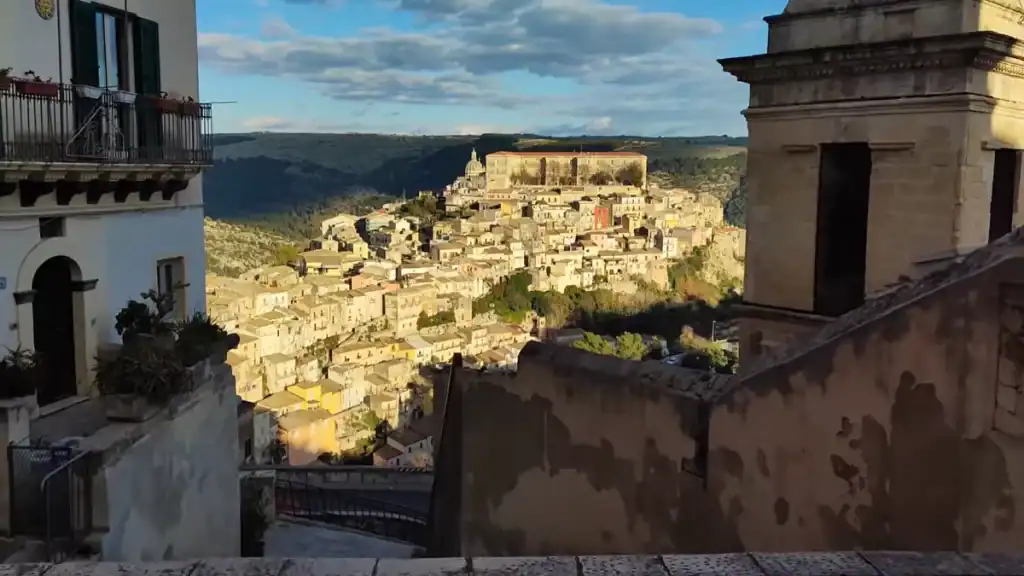 Ragusa Ibla spreads across its limestone ridge in warm evening light, seen through a gap between a baroque church tower and a residential building, with the deep Irminio valley gorge beyond.