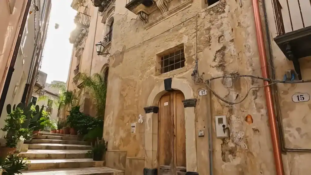 Worn limestone walls and a heavy wooden door at number 17 on a narrow stepped lane in Ragusa Ibla, potted plants crowding the staircase beside it