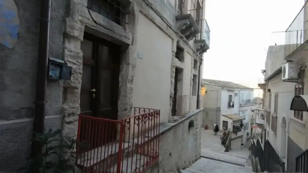 Narrow stepped street in Ragusa Sicily with a red iron gate in the foreground and limestone facades rising on both sides toward a small piazza below