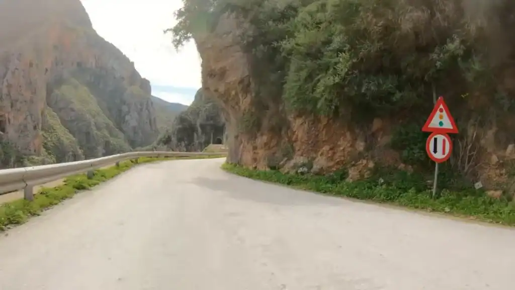 Single-lane mountain road threading through a steep gorge toward a tunnel entrance, Greek traffic signs warning of signals and narrow passage fixed to the rock face on the road to Elafonisi.