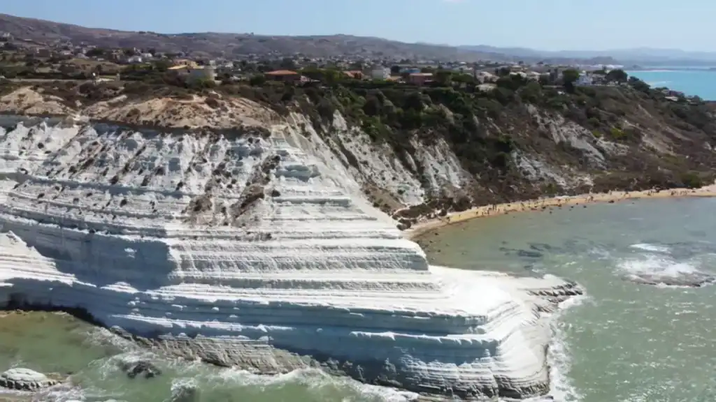 Layered white marl terraces of Scala dei Turchi dropping in broad natural steps to the sea, far from Ragusa Ibla, with a sandy beach curving beyond and a coastal settlement visible on the headland above.