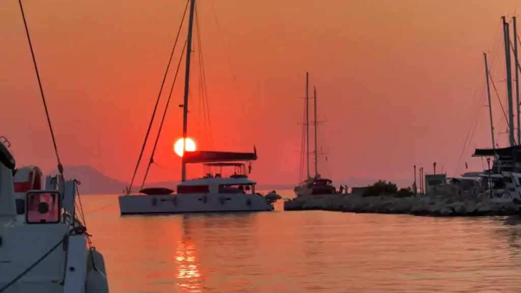 Catamaran silhouetted against a burning red sky as the sun sets directly behind its mast, moored boats and a stone breakwater visible to the right