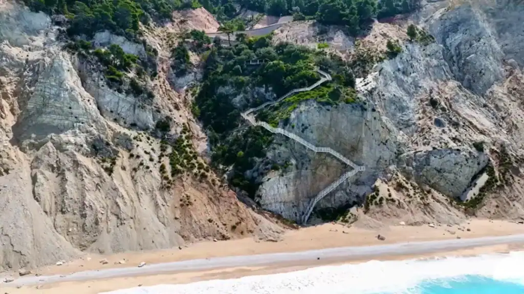 Zigzagging metal staircase cut into the face of Porto Katsiki's white cliff, descending in sharp switchbacks through sparse scrub to the sandy beach below.