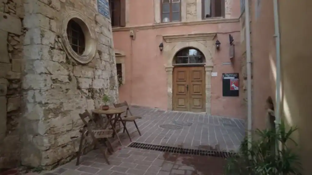A small wooden table and two chairs sit beside a rough stone wall in a quiet courtyard, facing a pink building with a arched wooden door