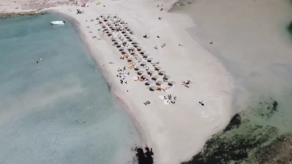 Aerial view of a narrow sandy spit at Balos Lagoon packed with sunloungers and parasols, water on both sides shifting from turquoise to pale green with bathers scattered across the sand