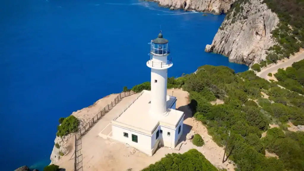Cylindrical white lighthouse with a green lantern room at Cape Lefkatas, its keeper's building set on bare rock surrounded by scrub, deep blue water and stratified cliffs visible behind.
