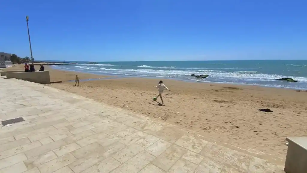 Child kicking a football on the sandy beach at Marina di Ragusa, with another child and two adults near a low wall beside the promenade, the harbour mole visible in the distance.