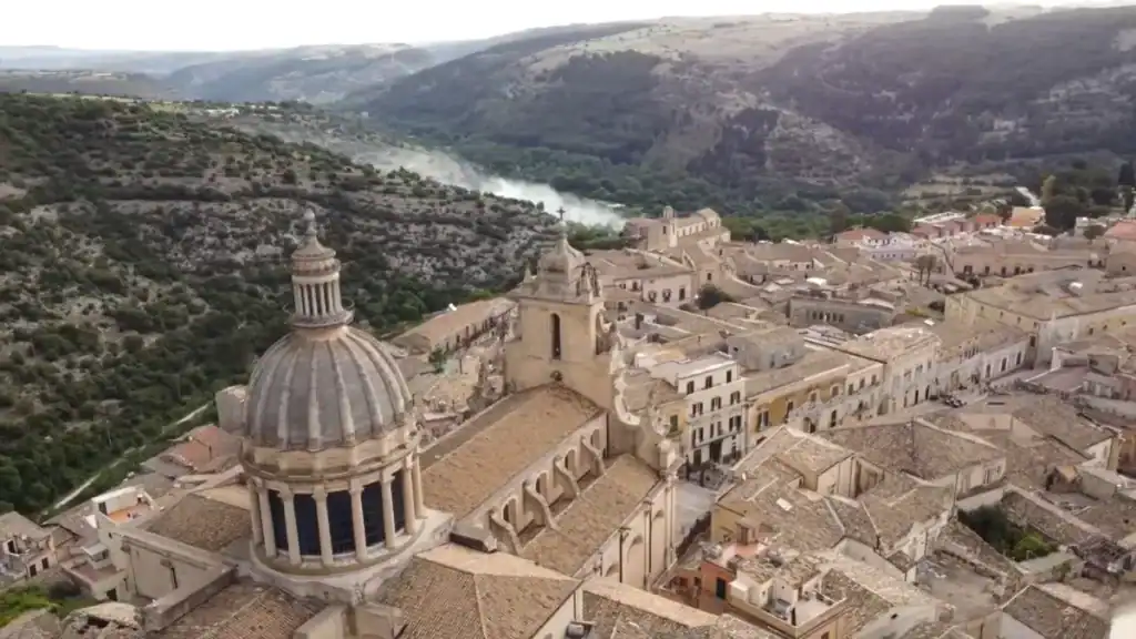Dome and bell tower of the Cathedral of San Giorgio rising above the limestone rooftops of Ragusa Ibla, wooded gorge visible behind