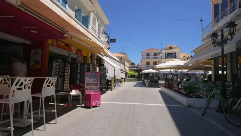 Cobbled pedestrian street lined with shop awnings, café seating under white parasols, and colourful two-storey buildings receding toward a small square.