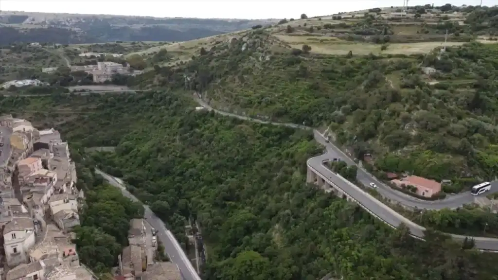 Aerial view of a switchback road descending into the Ragusa valley, dense tree canopy filling the gorge beside the old stone buildings of Ragusa Ibla