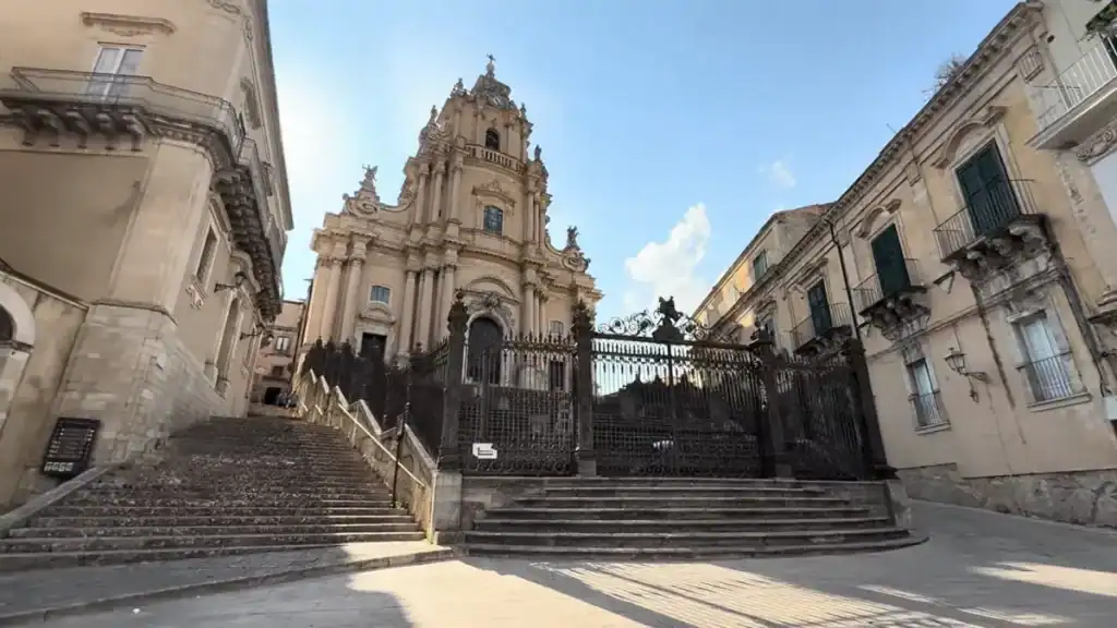 The Duomo di San Giorgio rises above wide stone steps and ornate iron gates, its layered baroque facade framing the Ragusa Ibla streets below
