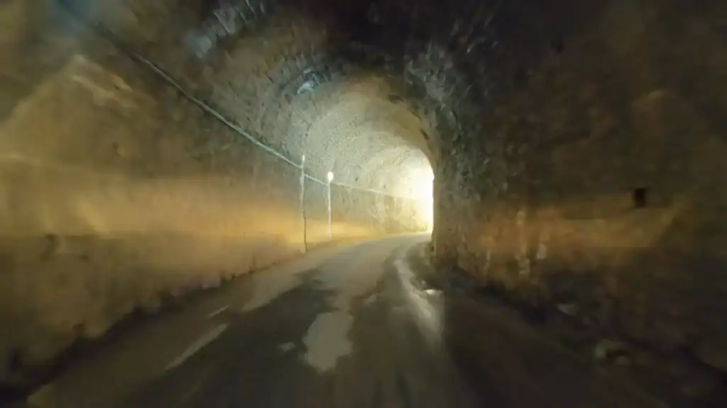 Inside a narrow stone-lined tunnel, rough brick arching overhead as the road curves toward a bright opening at the far end.