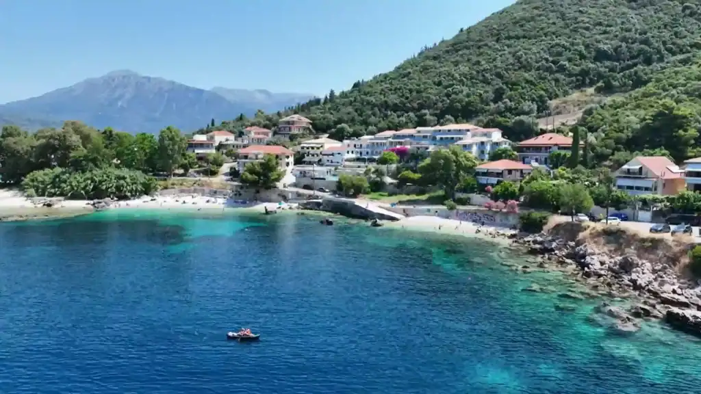 Vasiliki Lefkada from above, its horseshoe bay edged by two sandy beaches, terracotta-roofed buildings climbing into dense green hillside, a small motorboat crossing the deep blue foreground water.