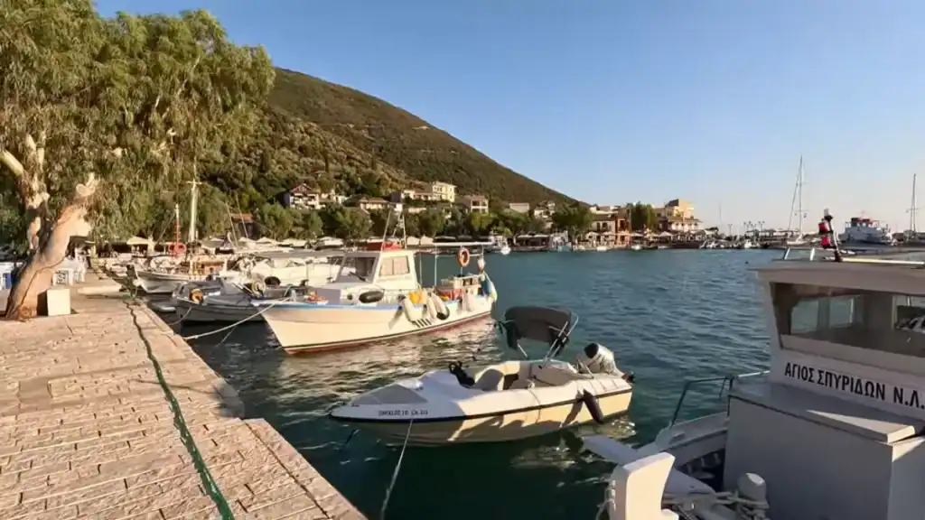 Fishing boats and small motorcraft moored along the stone harbour front, tavernas and a steep wooded hill visible across the water toward the village centre.