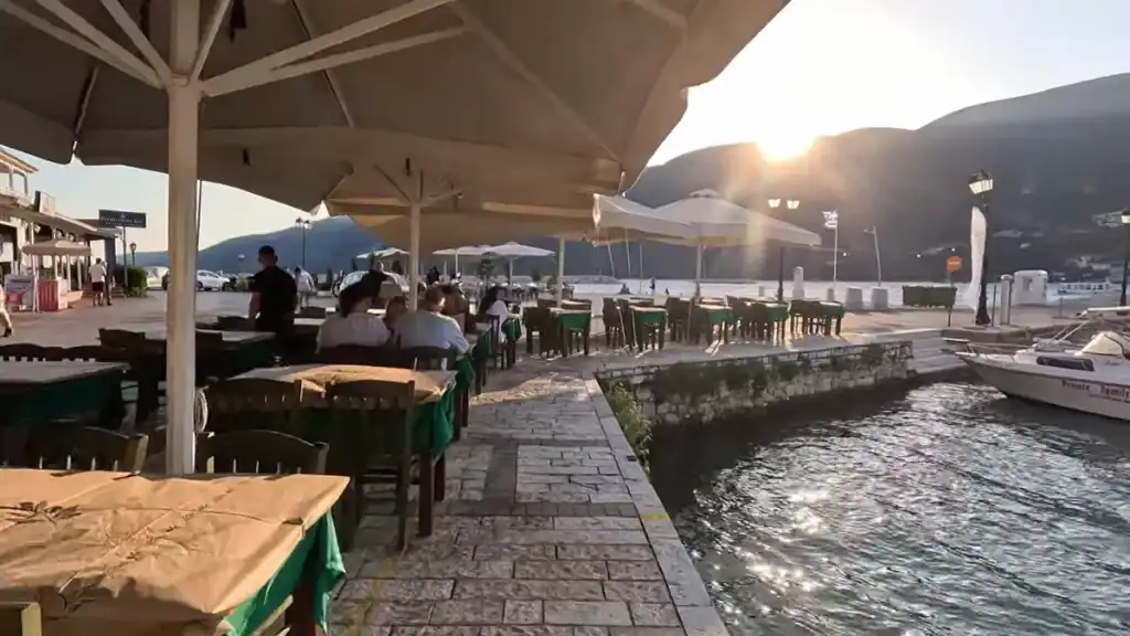 Diners seated under large parasols at a waterside taverna in Vasiliki Lefkada, the sun dropping behind the mountain ridge across the bay as a small boat sits moored at the quay edge.