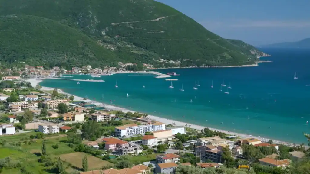 Vasiliki bay on Lefkada island, sailboats scattered across turquoise water in front of the village and its harbour, the settlement spreading along the shore beneath a steep green hillside.