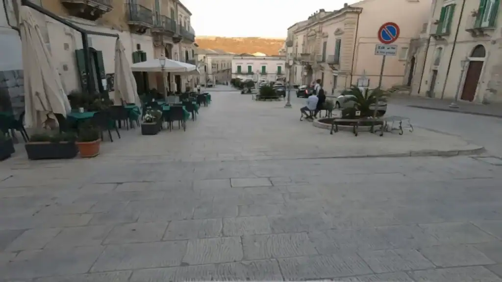 Quiet evening on Via Duomo in Ragusa Ibla, broad paved square with café tables set out on the left and two people seated at a central planter