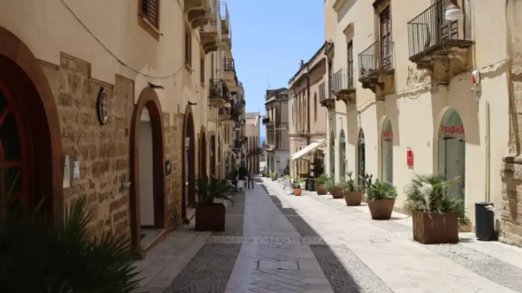 Narrow pedestrianised street lined with sandstone arcades, potted palms, and ornate baroque balconies receding toward a glimpse of blue sky at the far end.