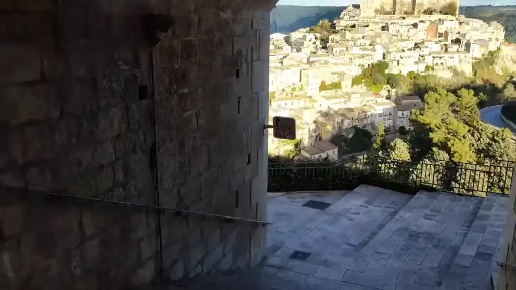 Stone wall of a building filling the left half of the frame, with Ragusa Ibla's white rooftops and a baroque church visible through the gap - part of walking in Ragusa along the stepped descent of Via Scale