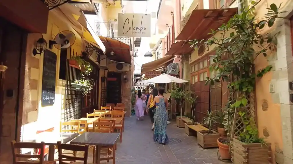 Narrow pedestrian lane lined with tavernas and cafe seating, people walking in Chania old town past the Colombo Kitchen Bar sign