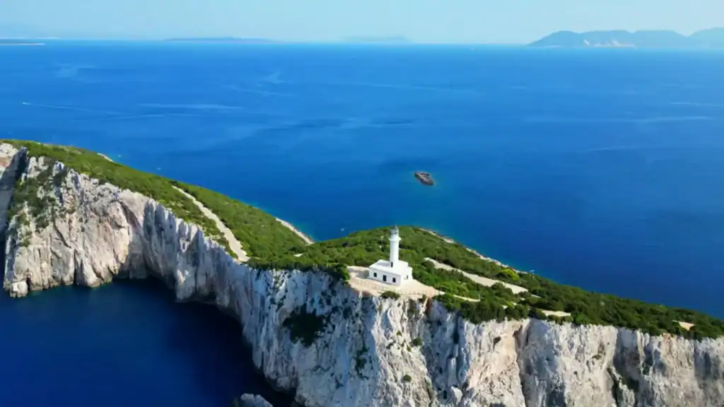 White lighthouse standing at Cape Lefkatas on sheer limestone cliffs above the Ionian Sea, with scrub-covered headland and distant islands visible across open water.