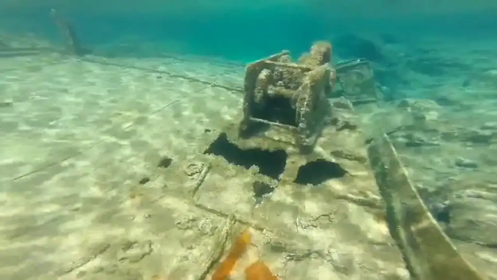 Encrusted metal machinery sitting on a flat submerged deck, planks and structural debris extending across a sandy seabed in shallow turquoise water.
