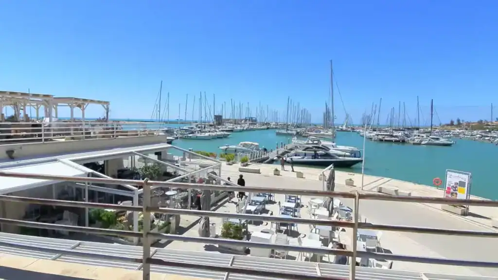 Waterside restaurant terrace overlooking a busy yacht harbour packed with sailboats, seen from an elevated position above the quayside.