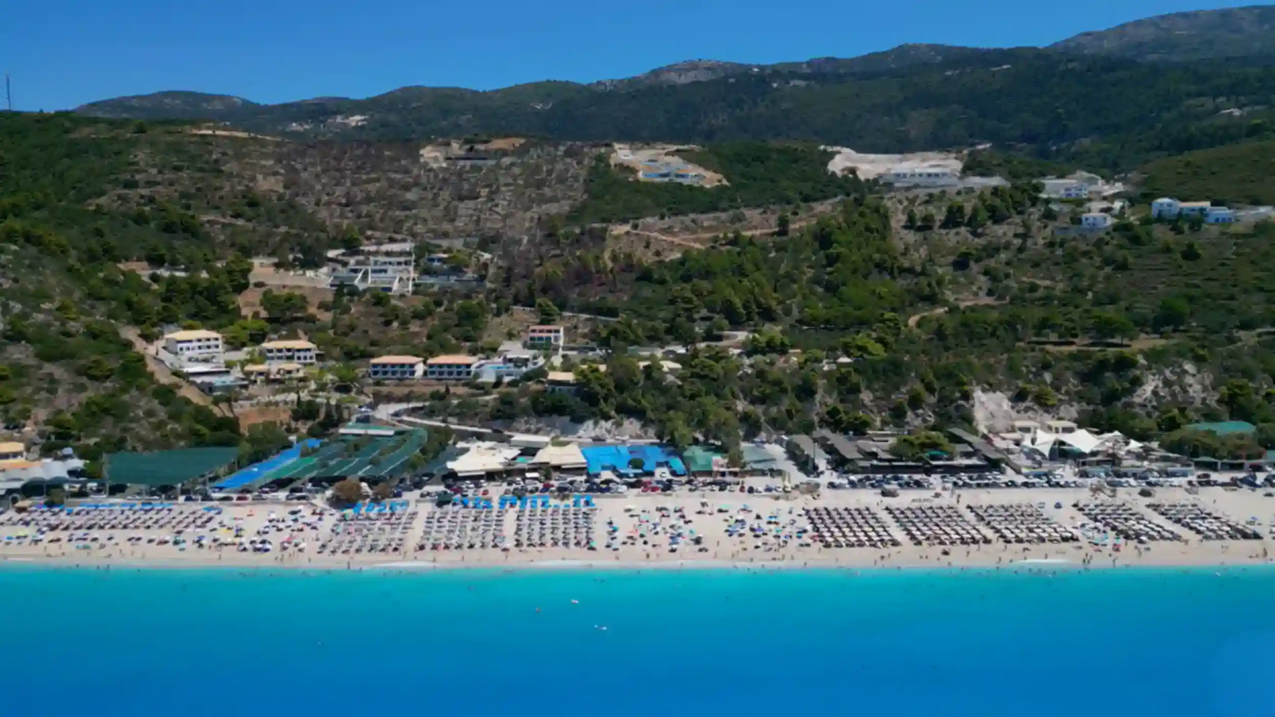 Kathisma beach from above, its long sandy shore packed with sunbeds and colourful umbrellas meeting vivid turquoise water, wooded hills rising steeply behind the seafront buildings