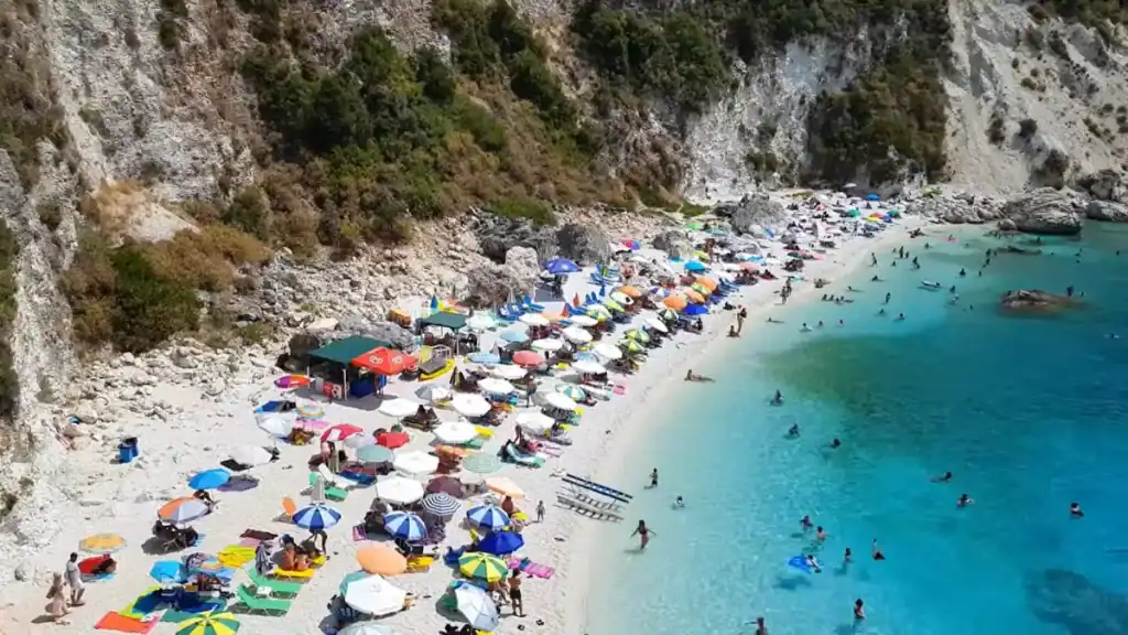 Hundreds of coloured umbrellas pack the full length of the pebble strip, a small beach bar visible mid-shore, swimmers spread across turquoise water with the enclosing cliff walls on two sides