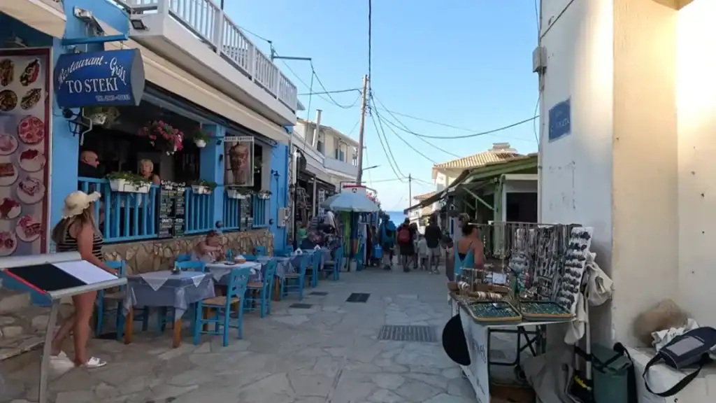 Pedestrian street in Agios Nikitas lined with tavernas and souvenir stalls, blue-painted restaurant chairs spilling onto the flagstones and the sea visible at the far end.