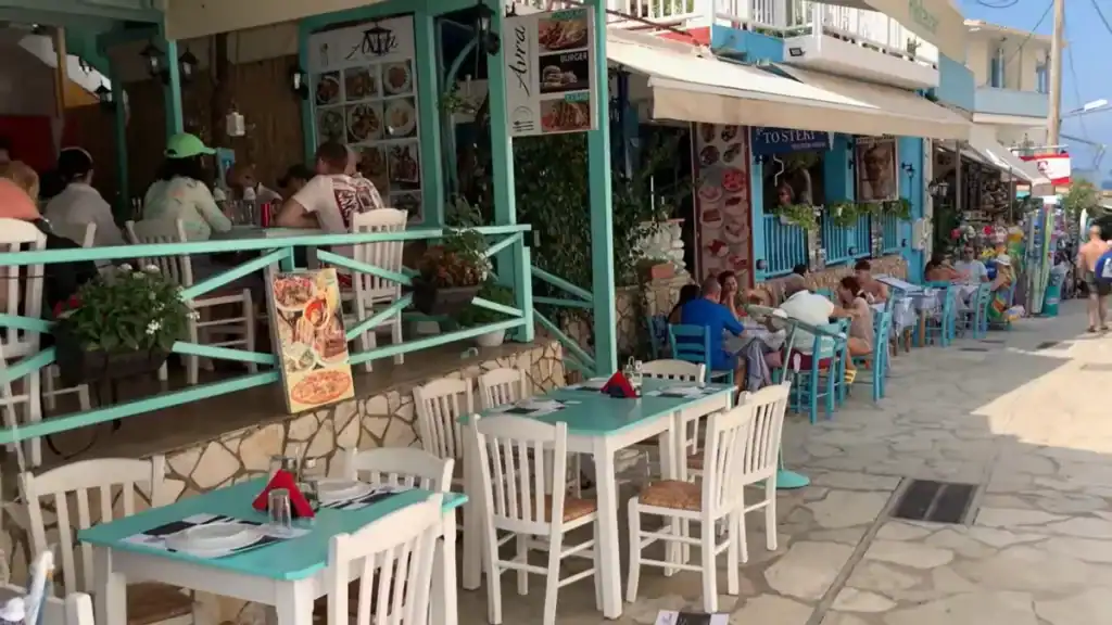 Outdoor seating spills onto a narrow pedestrian lane in Agios Nikitas, tavernas and casual eateries painted in turquoise and white stretching further down the street with diners already seated