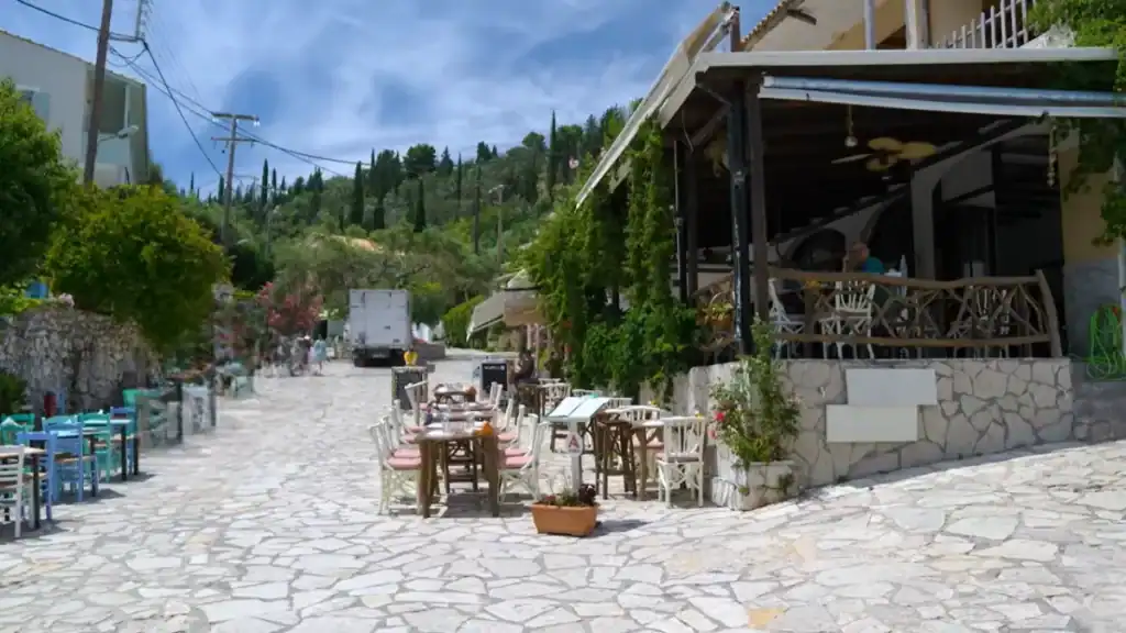 Taverna tables set out on a wide cobbled street in Agios Nikitas, vine-covered upper terrace to one side and wooded hills closing off the end of the village lane