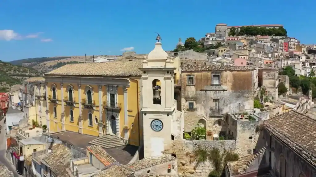 A bell tower with a clock face rises between a ochre-yellow baroque palazzo and weathered stone buildings in Ragusa Ibla, with the upper town stacked on the hillside behind.