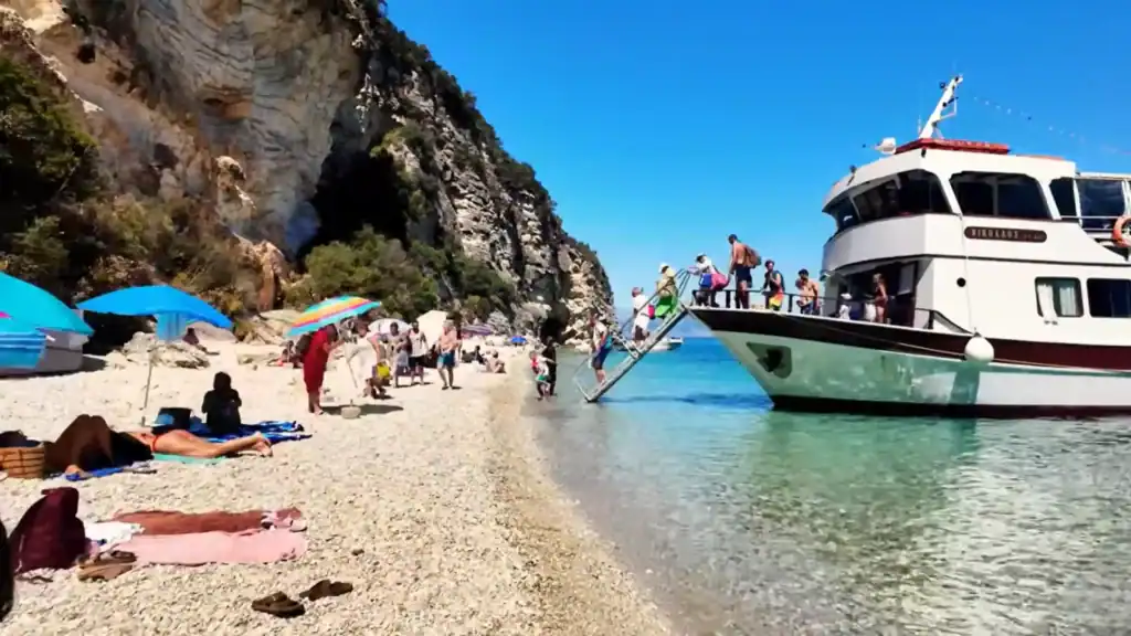 Passengers descend a gangway from a tour boat directly onto the pebbled shore at Agiofili Beach, sunbathers already settled under umbrellas as the limestone cliff walls the narrow cove behind