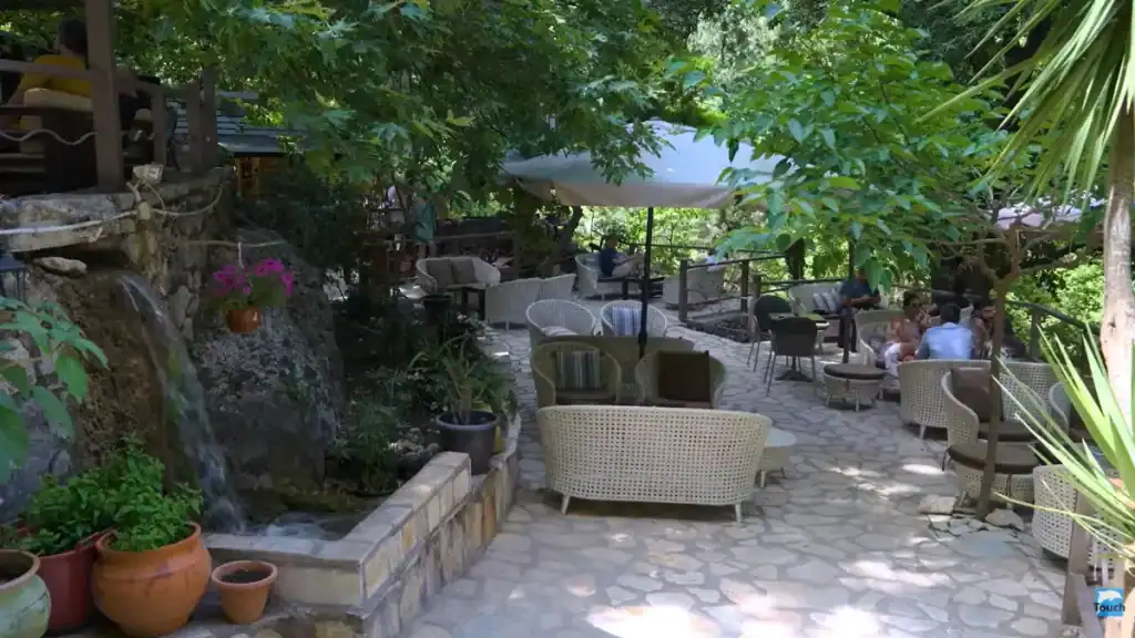 Wicker seating arranged across a stone-paved cafe terrace shaded by mature trees, a small waterfall trickling down a mossy rock face beside potted flowers at the edge of the patio