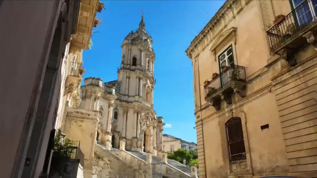 Ornate bell tower and facade of the Cathedral of San Giorgio seen from a narrow street below, flanked by weathered Baroque residential buildings with iron balconies - one of the finest examples of what draws visitors to Modica and Ragusa.