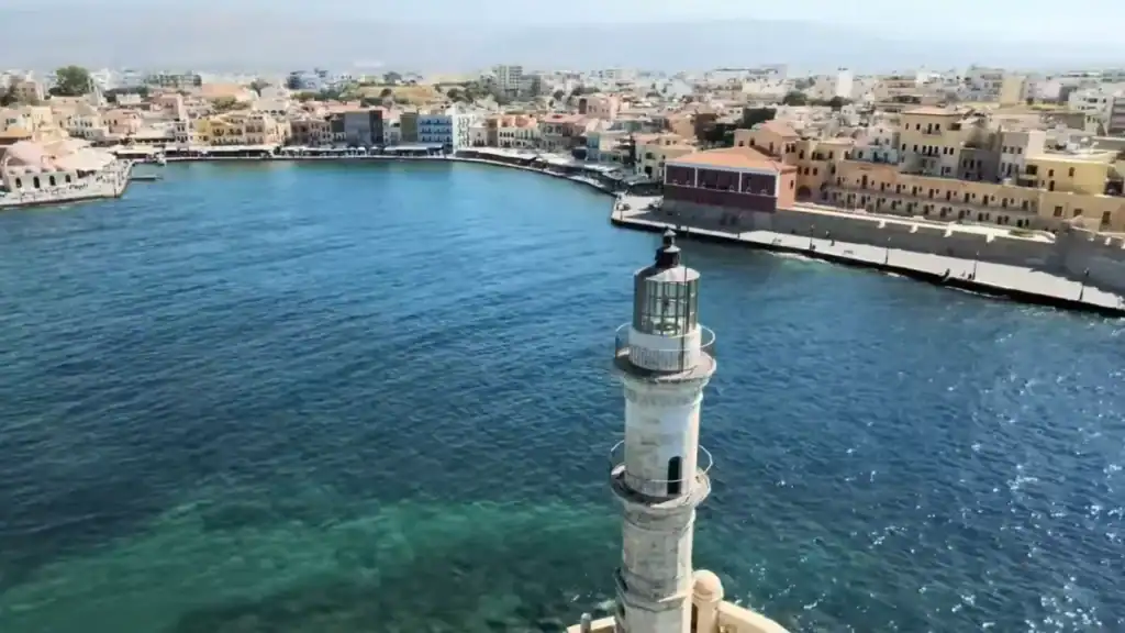 Chania lighthouse rising from the harbour entrance, the full arc of the old Venetian waterfront and city beyond spread across the frame