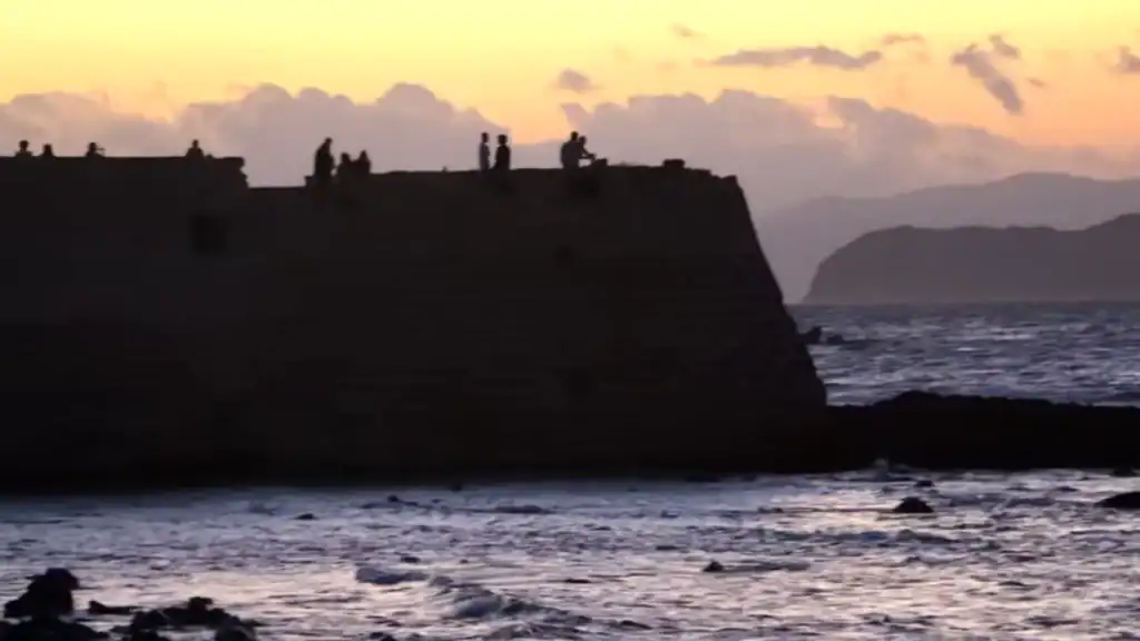 Silhouetted figures stand and sit along the top of the Firkas Fortress seawall at sunset, rough surf below and a headland fading into the orange-tinted dusk - one of those unhurried stops that defines solo travel in Chania.