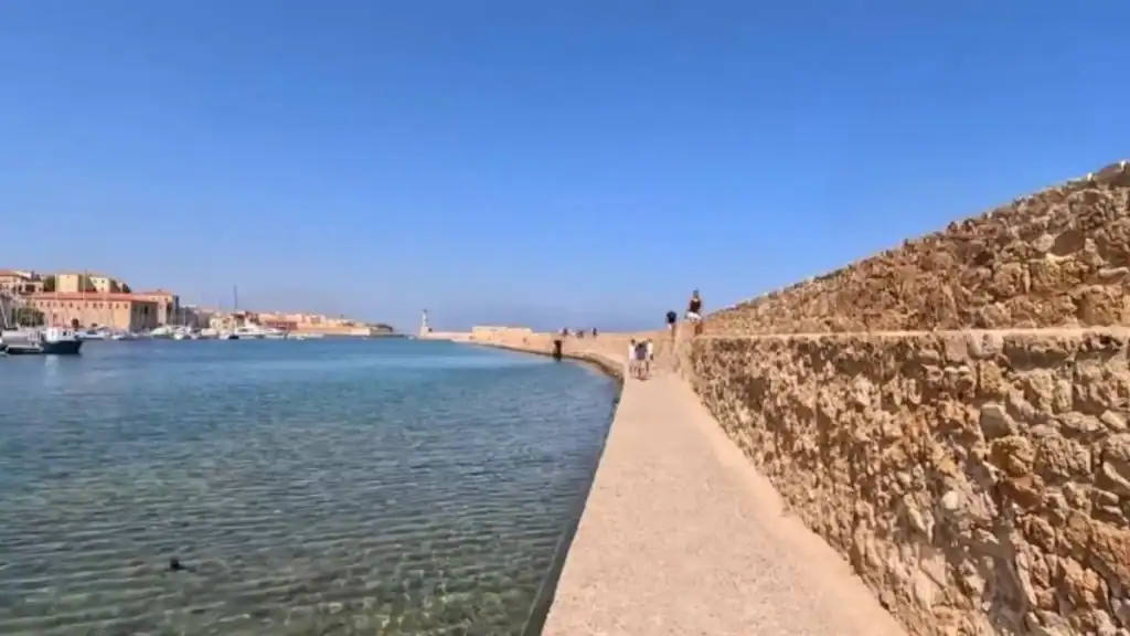 Stone breakwater walkway curving toward the Chania lighthouse, with the old harbour, moored boats and waterfront buildings stretching back to the left