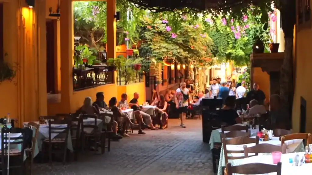 Diners at outdoor tables fill a vine-covered alley in Chania at dusk, warm lamplight catching amber walls as people move through - the kind of evening street that makes solo travel in Chania feel effortless.