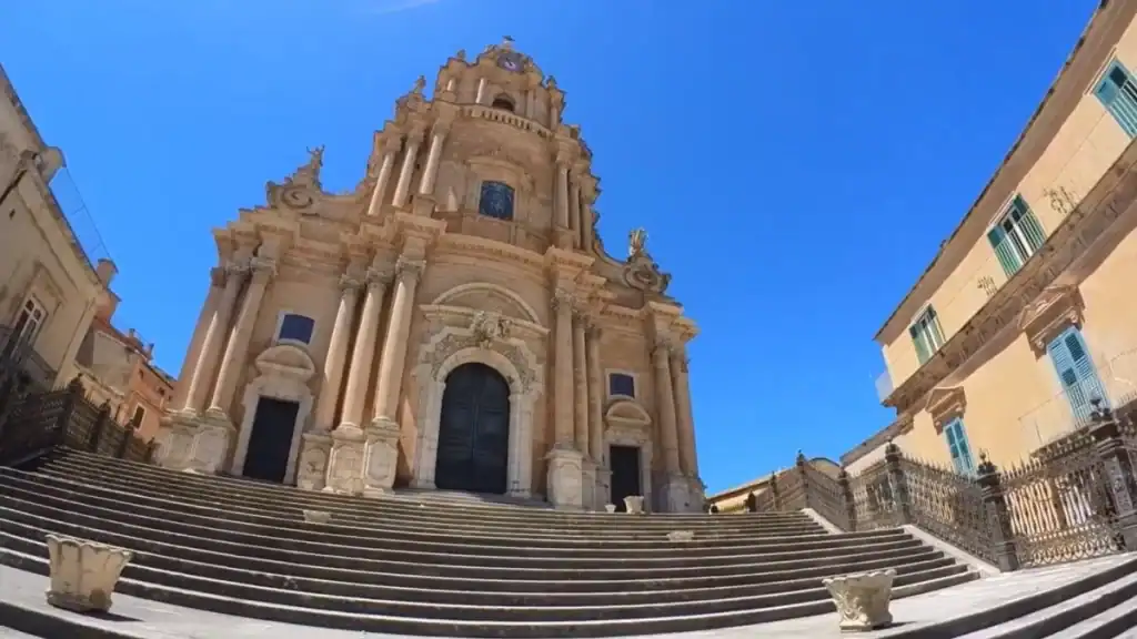 Wide stone staircase rising to the ornate Baroque facade of the Cathedral of San Giorgio in Ragusa Ibla, its tiered columns and sculptural detailing filling the frame from below.