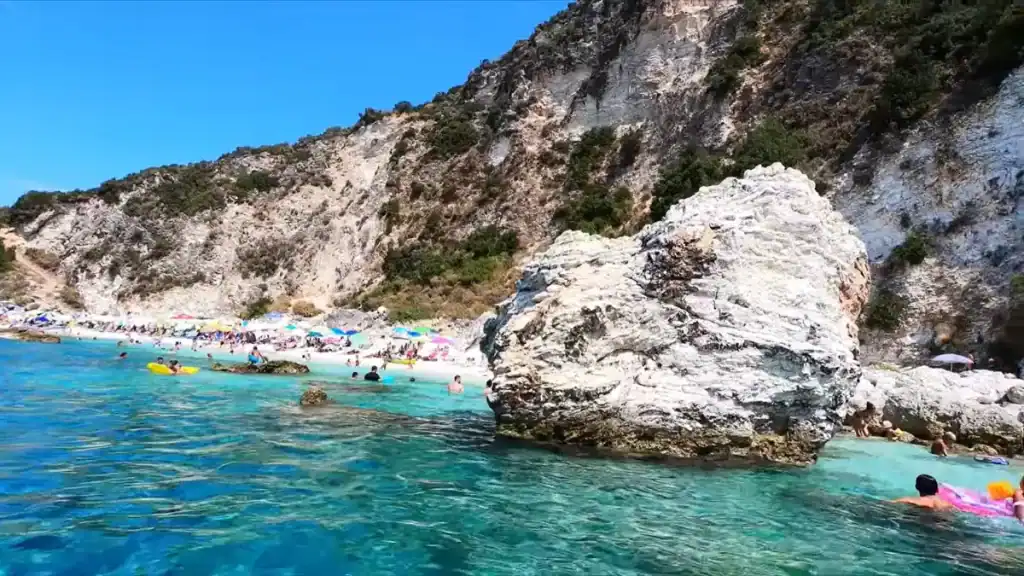 A large white limestone boulder rises from the water at the edge of a busy pebble beach, swimmers and inflatable floats scattered across the shallows with the full cliff face stretching behind