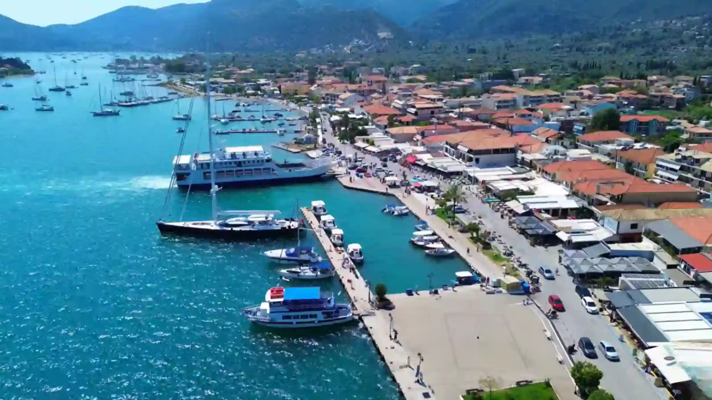 Aerial view of Nidri Lefkada harbour with a large ferry docked along the main quay, smaller boats moored at a central pier, and the village seafront road lined with shops and terracotta-roofed buildings backed by forested hills
